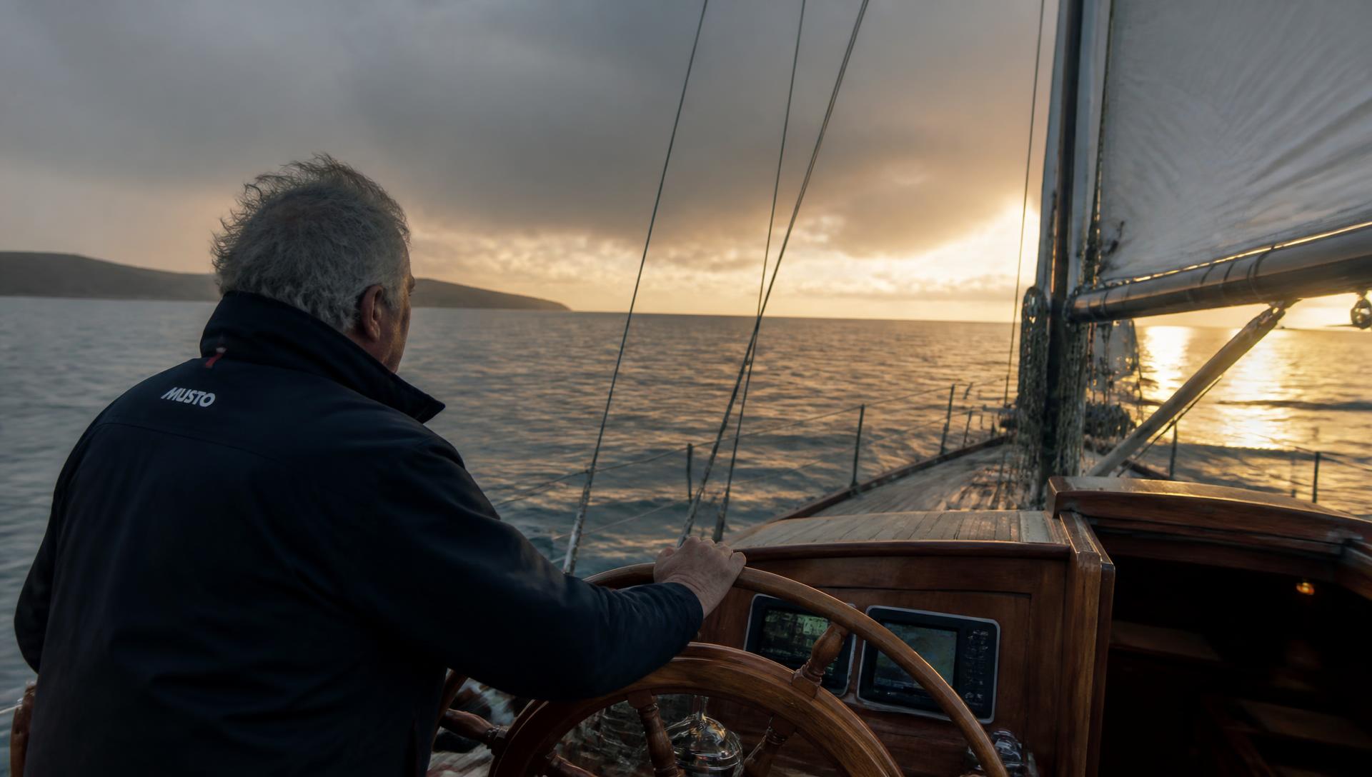 An experienced British skipper at the wooden helm of a classic sailing yacht, steering towards a bright horizon on calm water after rough weather